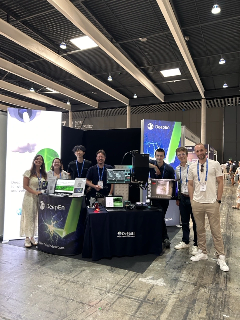 Seven DeepEn team members standing at the company’s FENS 2026 booth beside the DeepEn One microendoscopy demonstration system.